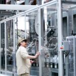 Man observes and operates advanced industrial machinery behind glass indoors.